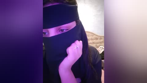 Media: Video of a person in black mask, head tilted, hand on cheek, eyes visible, against a blurred background with a beige headboard.