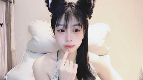 Media: Video of a young Asian woman with long black hair, wearing black cat ears and a white lace dress, resting on a white cushioned chair against a beige wall.