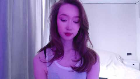 Media: Video of a slender young woman with long brown hair, fair skin, and red lipstick, wearing a white tank top, standing in a softly lit, minimalistic bedroom with white walls and a bed.