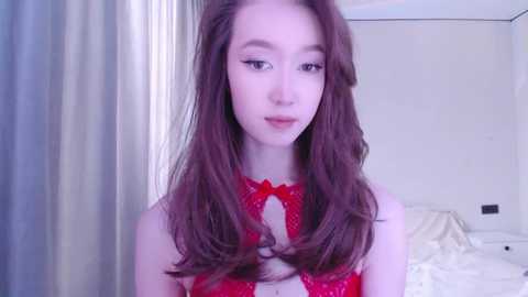 Media: Video of a young, fair-skinned woman with long, wavy brown hair, wearing a red lace bralette, standing in a softly lit bedroom with white curtains and a bed in the background.