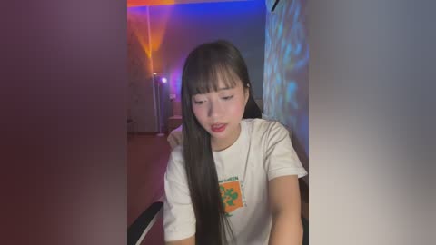 Media: Video of an Asian woman with long black hair, wearing a white t-shirt with a green emblem, sitting indoors with colorful LED lights in the background.