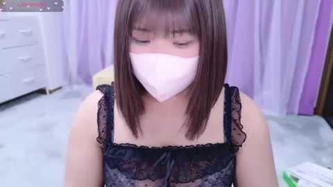 Media: Video of a young Asian woman with straight brown hair and light skin, wearing a black lace lingerie top and white face mask, seated indoors with white drawers and lavender curtains in the background.