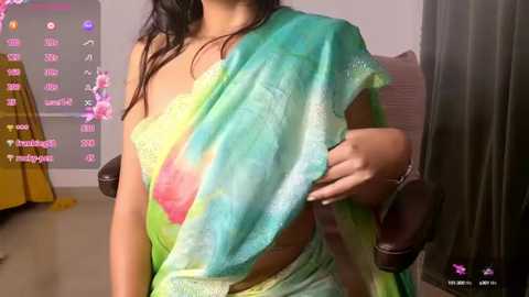 Media: Video of a woman with medium brown skin and long black hair, wearing a colorful, semi-transparent saree. She is sitting on a brown chair, holding her left breast. Background shows a yellow bed, a window, and a gray curtain.