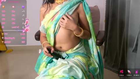 Media: Video of a topless woman in a green and yellow saree, covering her breasts with one hand, wearing gold jewelry. Background includes a gray wall and a wooden chair.