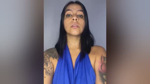 Media: Video of a young woman with medium brown skin, long black hair, and tattoos on her arms, wearing a blue halter top, standing against a blurred, neutral background.