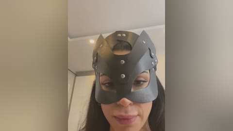 Media: Video of a woman with long dark hair, wearing a gray, cat-like mask with two small holes and silver studs. The background shows a white ceiling and a door frame.