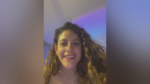 Media: Video of a smiling, curly-haired woman with light skin, wearing a white top, in a dimly lit room with purple and blue lighting.