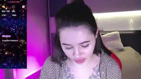 Media: Video of a young woman with light skin and dark hair, wearing a black and white patterned top, looking down, in a bedroom with a nightstand and bed.