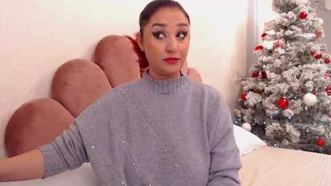 Media: Video of a woman with dark hair in a ponytail, wearing a light gray sweater, seated on a bed with a brown headboard and snow-covered Christmas tree in the background.