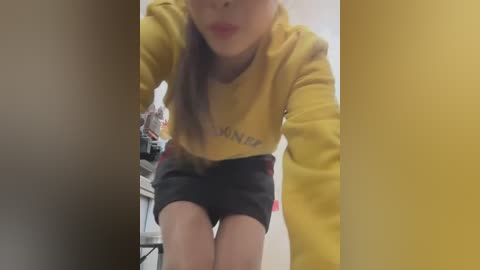 Media: Video of a young woman with light skin, wearing a yellow sweatshirt and black shorts, leaning forward, lips slightly parted, in a dimly lit room with beige walls.