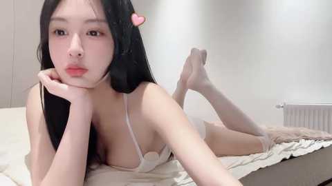 Media: Video of a young East Asian woman with long black hair, wearing a white bra, lying on a beige bed, her legs raised, with a pink heart hair clip.