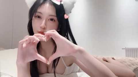 Media: A video of an Asian woman with long black hair, wearing a white lingerie top, posing in a heart shape with her hands in a minimalist bedroom with white walls and a radiator.