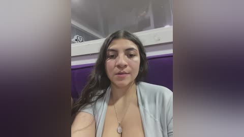 Media: A video of a Latina woman with long dark hair, medium skin tone, and a light gray cardigan, sitting indoors against a purple wall. Her expression is neutral, and she wears a necklace.