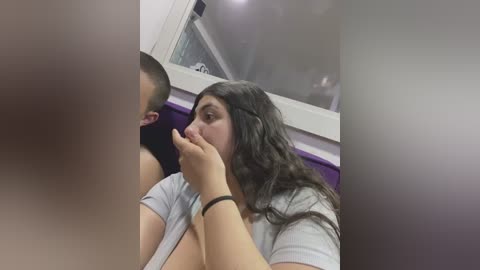 Media: A video of a young woman with long, dark hair in a grey shirt, covering her mouth with her hand, sitting in a public bathroom stall. The background shows a purple wall and a window.