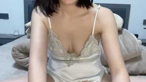 Media: A video of an Asian woman with shoulder-length black hair, wearing a revealing, beige satin camisole with lace trim, sitting on a bed with beige bedding. The room is softly lit with a neutral color palette.