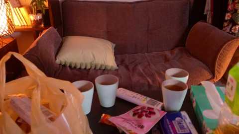 Media: Video of a cozy, dimly-lit living room with a beige couch, a beige pillow, two white cups of tea, a beige shopping bag, and various snack boxes scattered on a dark wooden table.