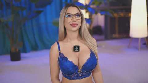 Media: Video of a blonde woman with glasses, wearing a blue lace bra, standing indoors against a backdrop of green plants and a white lamp.