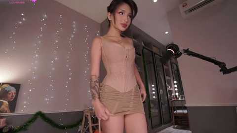 Media: Video of a slender Asian woman with fair skin, wearing a tight, beige corset, short skirt, and dramatic makeup, standing in a well-lit, modern room with fairy lights, a large mirror, and studio lights.