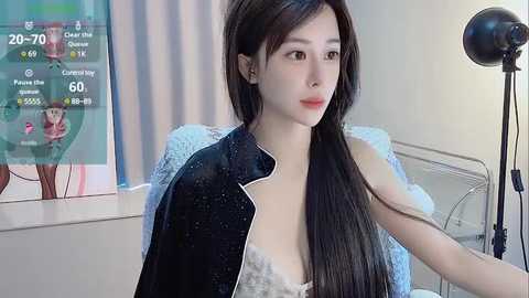 Media: Video of a young Asian woman with long black hair, fair skin, and large eyes, wearing a black robe, sitting in a modern, bright room with a large screen displaying game stats.