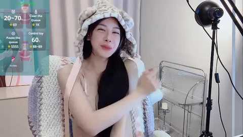 Media: A video of an East Asian woman with long black hair and fair skin, wearing a cozy, knit hat and robe, smiling in a brightly lit room with a studio light and a birdcage.