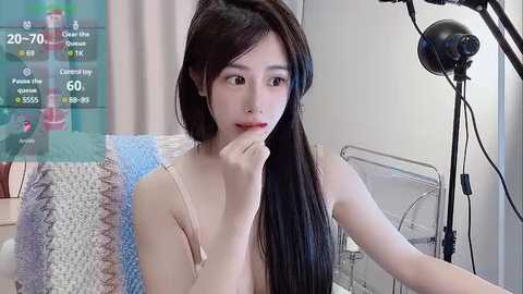 Media: A video of an East Asian woman with long, straight black hair, fair skin, and a slender physique, sitting on a couch. She wears a beige tank top, with a transparent overlay showing a live stream screen.