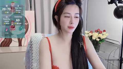 Media: Video of an East Asian woman with long black hair, wearing a red headband and a red bra, sitting in a room with a grey chair, a vase of flowers, and a wall-mounted spotlight.