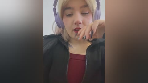 Media: Video of a young woman with fair skin, blonde hair, wearing purple headphones, black jacket, and red top, biting her finger in a contemplative pose.