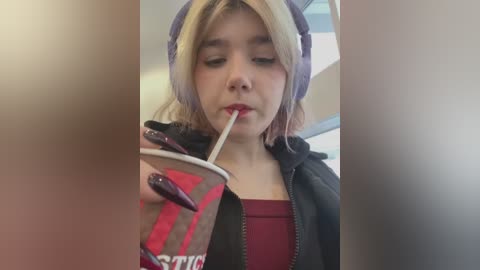 Media: Video of a young girl with light skin, blonde hair, and large headphones, drinking from a red and white striped cup, wearing a maroon top and black jacket, indoors.