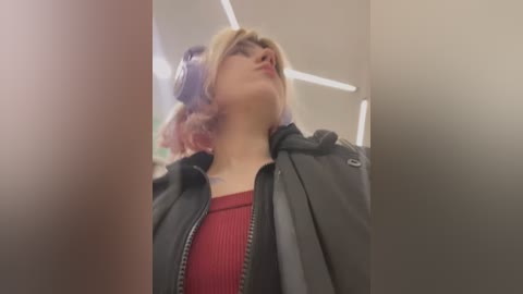 Media: A video of a young woman with pale skin and pink hair, wearing black headphones, a red sweater, and a black jacket, looking upward. The background features a modern, indoor setting with white ceiling lights.