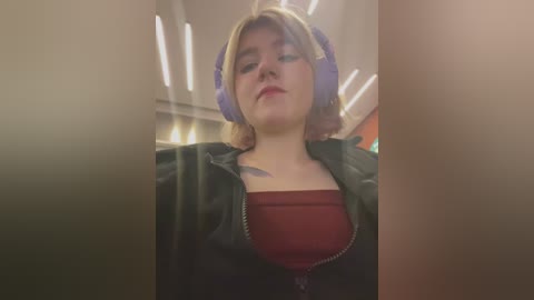 Media: A video of a young woman with short, blonde hair wearing purple headphones and a black jacket over a red shirt, sitting in a dimly lit, modern interior.