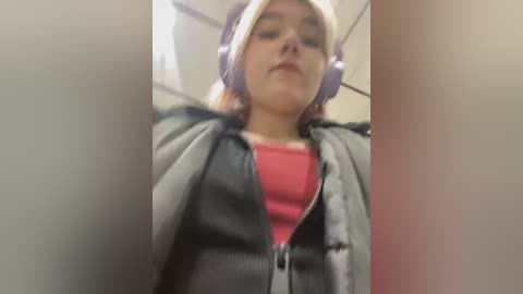 Media: A blurry video of a young woman with light skin, wearing a pink top, gray jacket, and purple headphones, standing in an indoor setting with a beige background.