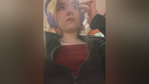 Media: Video of a young woman with light skin and blonde hair, wearing purple headphones and a black jacket over a red shirt, adjusting her glasses.