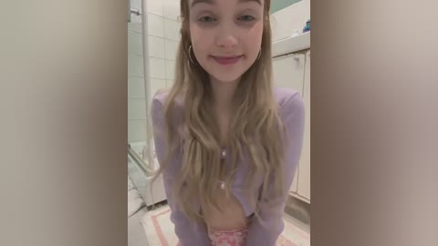 Media: Video of a young woman with long blonde hair, wearing a lavender cardigan and pink pajama pants, smiling, in a bathroom with white tiles and a white sink.