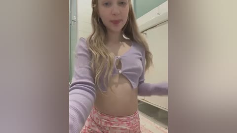 Media: Video of a young Caucasian woman with long, wavy blonde hair, wearing a purple cropped top and red patterned shorts. She's standing in a bathroom with beige walls and a sink.