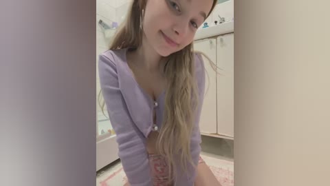 Media: A video of a young Caucasian woman with long, wavy blonde hair, wearing a lavender cardigan, kneeling on a pink rug in a bathroom with white cabinets and a bathtub.