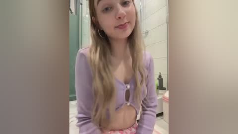 Media: Video of a young Caucasian woman with long blonde hair, wearing a cropped lavender cardigan and pink shorts, sitting in a bathroom with white tiles and a pink soap dispenser.