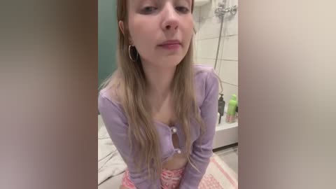 Media: Video of a young Caucasian woman with fair skin and long blonde hair, wearing a purple crop top and patterned shorts, sitting on a bathroom floor. Background shows a white-tiled shower area with toiletries.