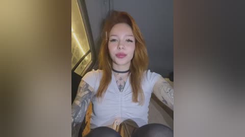 Media: Video of a tattooed, pale-skinned woman with long red hair, wearing a white shirt and black choker, making a pouty face in a dimly lit room.