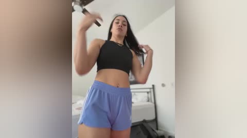 Media: Video of a fit woman with long dark hair, medium skin tone, wearing a black sports bra and blue shorts, flexing her arms in a bedroom with a bed and white walls.