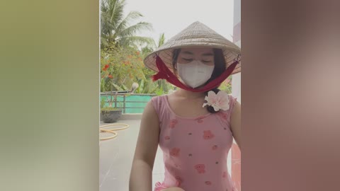 Media: Video of a young Vietnamese woman in a pink, floral-patterned shirt and conical hat, wearing a face mask, standing outdoors with a green fence and palm trees in the background.