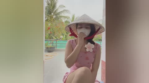 Media: A video of an Asian woman wearing a conical hat, pink dress, and mask, sitting on a boat, with a tropical background of palm trees and greenery.