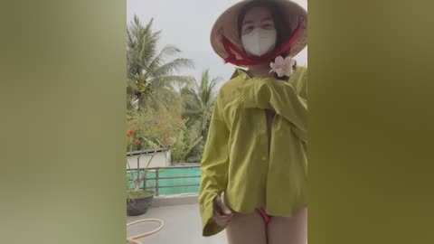 Media: Video of an Asian woman in a green shirt, red thong, and wide-brimmed hat, standing outdoors with a mask on, surrounded by lush greenery and a pool.