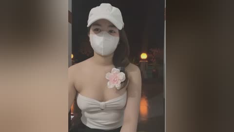 Media: Video of a young Asian woman with light skin, wearing a white face mask, a white cap, and a strapless top with a pink flower brooch. She stands indoors, with a blurred background featuring dim lighting and indistinct shapes.