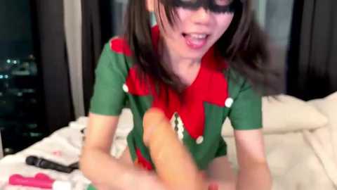 Media: A video of an Asian woman with straight black hair and a green and red elf costume, licking a large dildo, set against a dark backdrop with a bed.