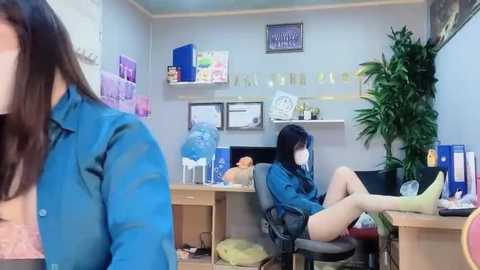 Media: A video of an office room with a woman in a blue shirt and white mask sitting on a desk, another woman in a blue shirt in the foreground, and a green plant in the background.