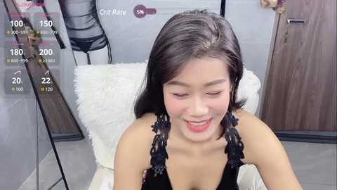 Media: Video of an Asian woman with long black hair, wearing a black lace top, sitting on a white chair in a modern room.