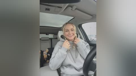 Media: Video of a smiling, blonde woman wearing a gray hoodie, sitting in a car with a child's stuffed animal and a snack bag in the background.