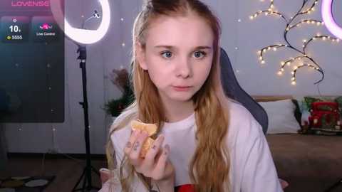 Media: A video of a young girl with long blonde hair, wearing a white shirt, eating a sandwich, in a festive, dimly-lit room with string lights and a ring light.