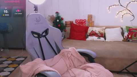 Media: A video of a cozy living room featuring a light grey gaming chair with a bow design, a beige couch with festive pillows, a lit Christmas tree, and a soft pink blanket.