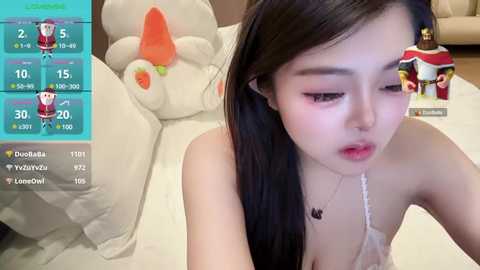 Media: Video of a young Asian woman with long black hair, wearing a white lace bra, sitting on a plush toy bed, in a cozy room with plush toys and a TV screen displaying game stats.